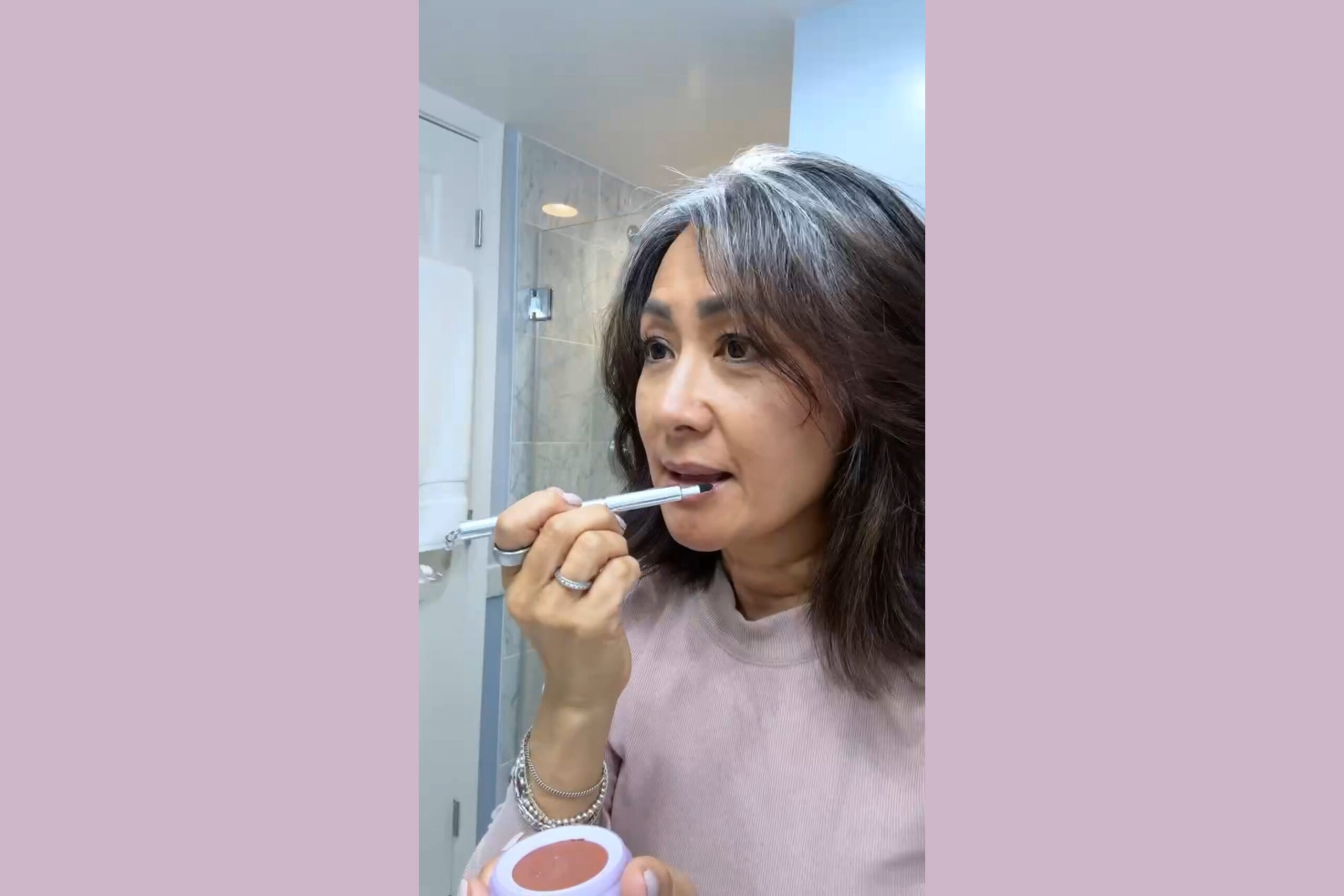 Me applying makeup in a hotel bathroom during a trip—visible natural gray roots, no dye, and a calm, confident expression. A moment of freedom in my gray hair era.