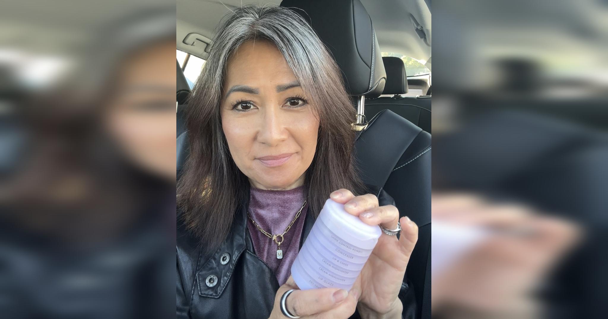 Midlife woman with natural gray hair holding Subtl Beauty Stak in car, wearing soft makeup and a leather jacket.