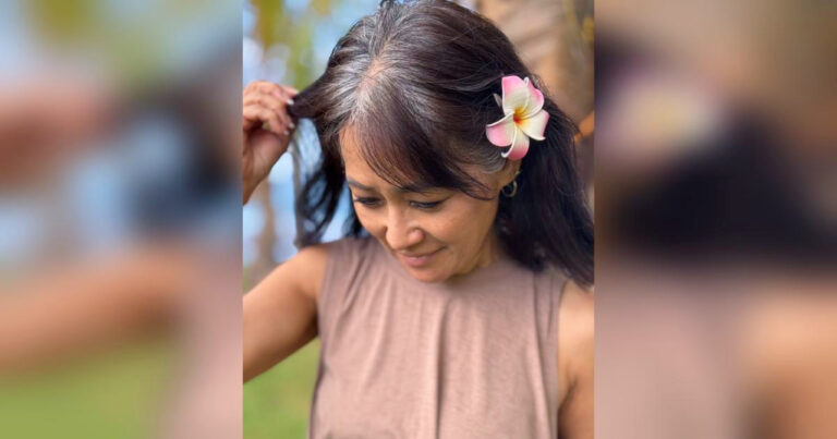 Midlife woman beginning her natural gray hair transition, gently parting her hair to reveal early silver strands, wearing a plumeria flower and soft expression.