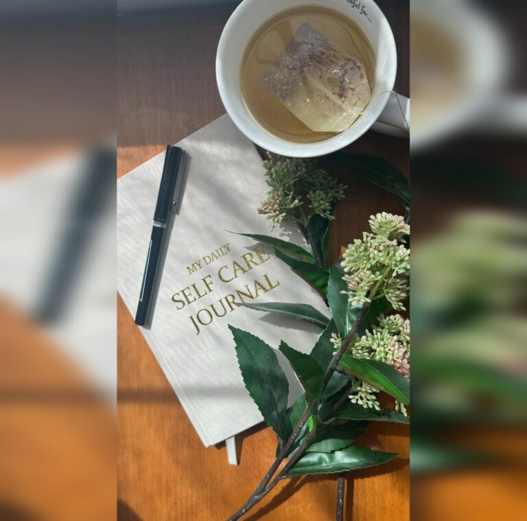 Flat lay of a self-care journal, pen, herbal tea, and floral sprig in soft natural light—evoking calm, reflection, and mindful beginnings.