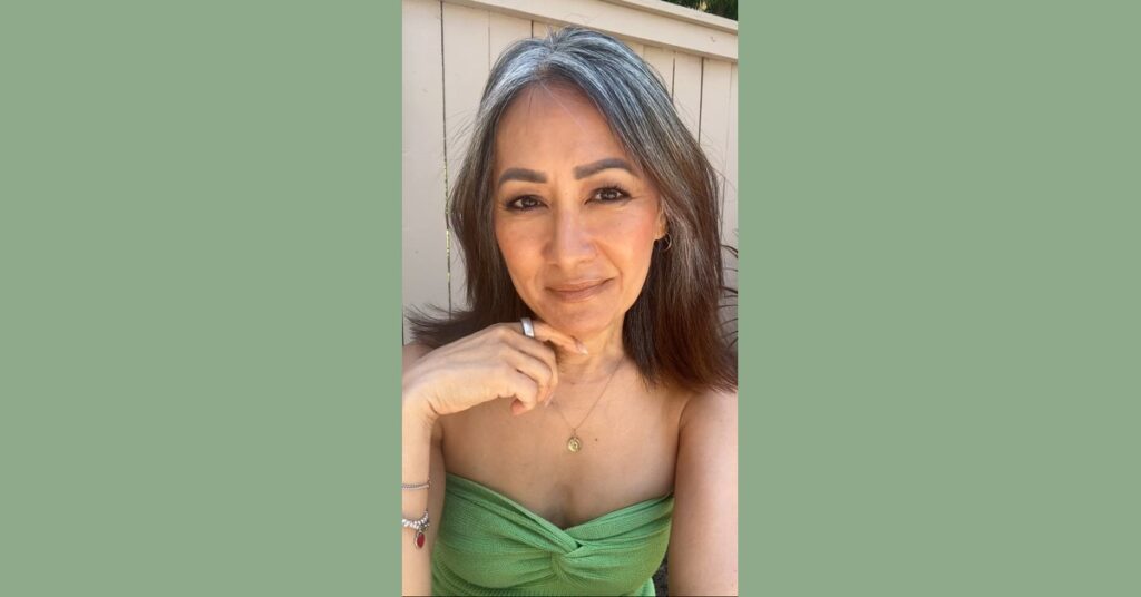 Woman with natural gray streaks and medium-length dark hair, smiling in sunlight wearing a strapless green knit top.