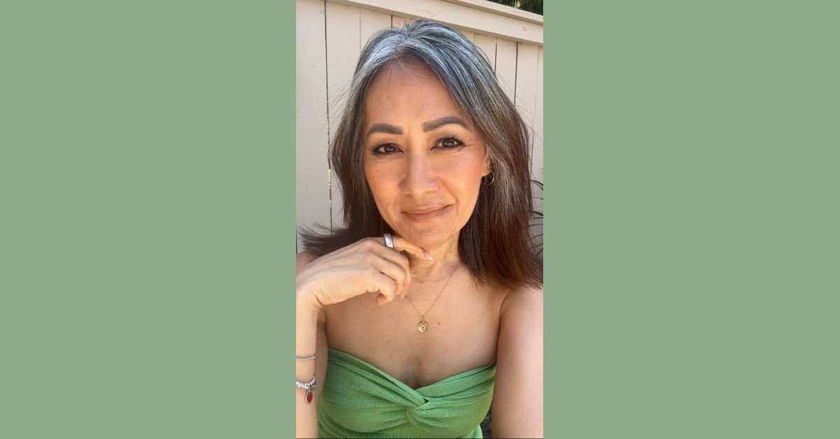 Woman with natural gray streaks and medium-length dark hair, smiling in sunlight wearing a strapless green knit top.