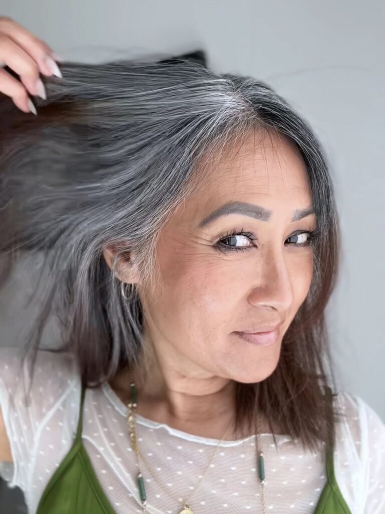 1. Meesh holding up one side of her hair to show the blend of silver and dark strands at nine months into her gray grow-out journey, smiling with soft confidence.
2. Back view of Meesh’s hair showing a natural V-shaped pattern of gray growth blending into the darker ends—highlighting the unique and personal nature of her grow-out.
3. Front-facing portrait of Meesh at nine months into her gray hair transition, smiling with her blended gray and brunette strands softly framing her face.
