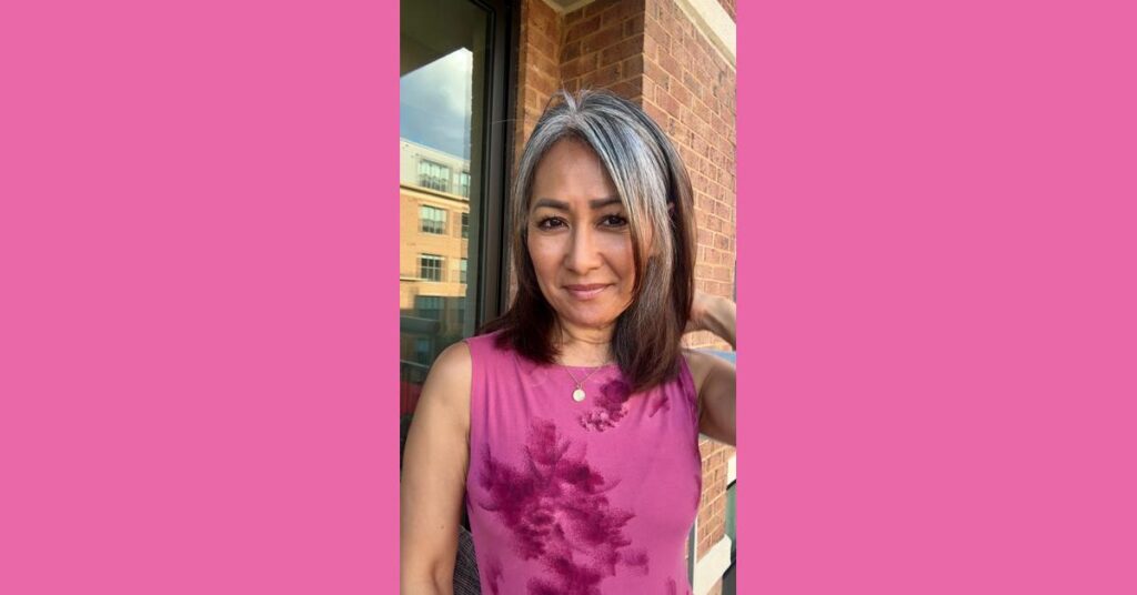 Midlife woman embracing her gray hair journey, smiling in a magenta floral top against a brick wall.