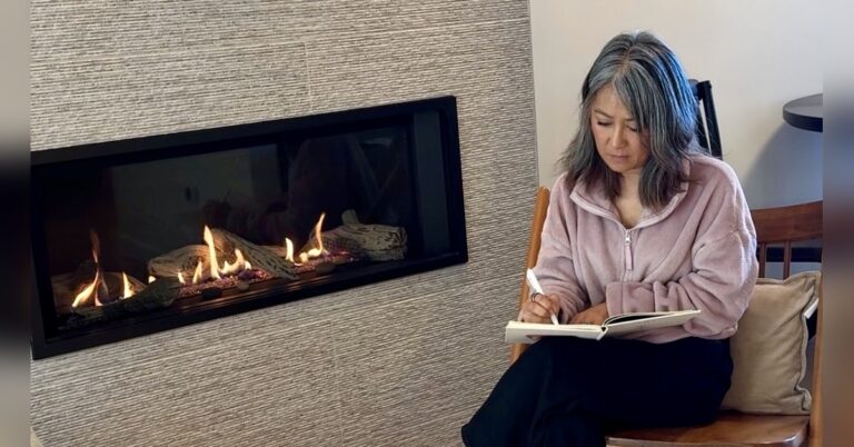 Woman with gray hair sitting by a fireplace and writing in a journal in a calm, reflective space.