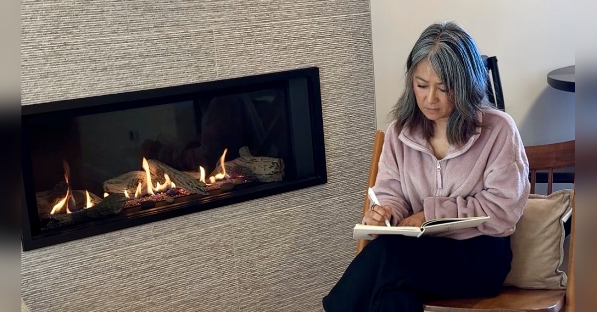 Woman with gray hair sitting by a fireplace and writing in a journal in a calm, reflective space.